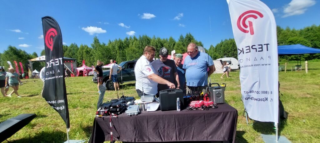 Group of men at an outdoor tech demonstration table, inspecting radio equipment on a sunny day.
