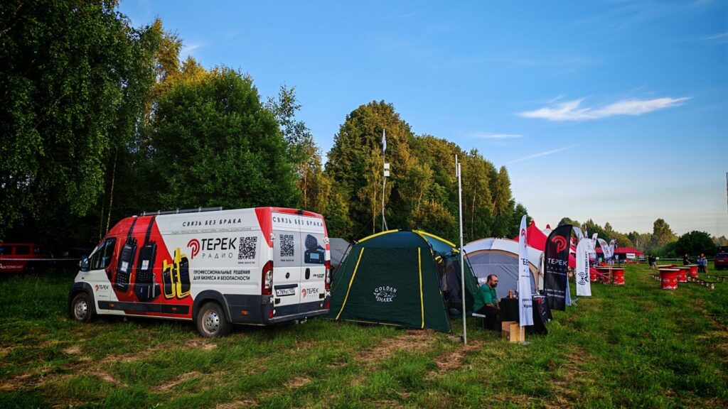 Red and white promotional van with Cyrillic text parked beside green tents at an outdoor festival, with trees in the background under a blue sky.