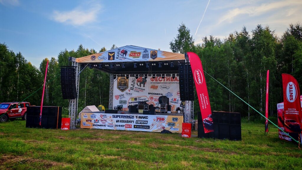 Outdoor festival stage with sponsor banners and tall red flags, set in a grassy clearing by a forest.