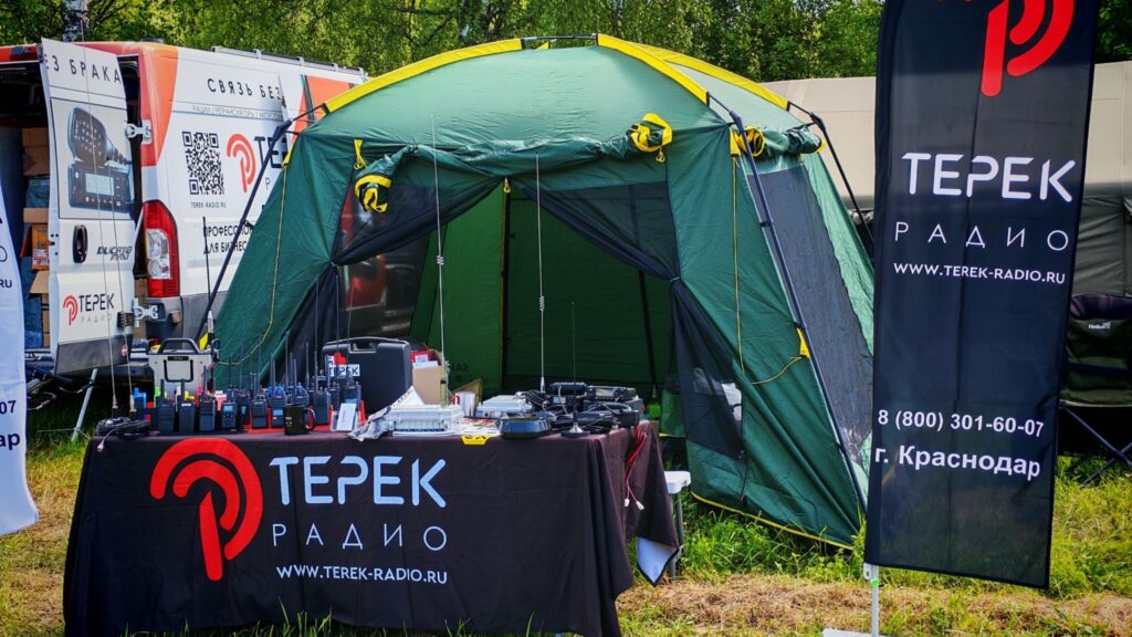 Outdoor TEREK Radio promotional booth with a green tent, tables full of radios and equipment, and a tall banner reading 'ТЕРЕК РАДИО'.
